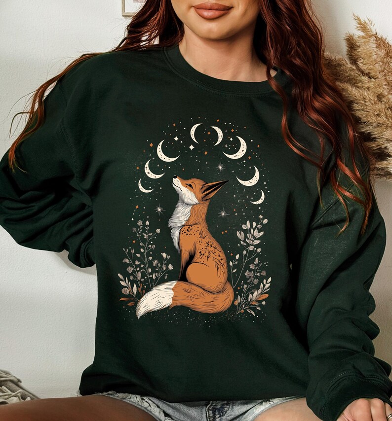 K&ouml;nnte beinhalten: Dunkelgr&uuml;nes Sweatshirt mit einer detaillierten Illustration eines Fuchses, der auf ein Mond- und Sternenmuster blickt. Der Fuchs ist braun und wei&szlig;, umgeben von floralen Elementen. Das Design ist mittig auf dem Sweatshirt platziert.
