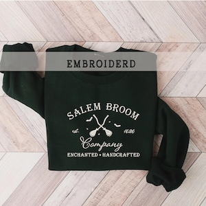 K&ouml;nnte beinhalten: Ein schwarzer Pullover mit wei&szlig; gesticktem Text, der "Salem Broom Company est. 1626 Enchanted + Handcrafted" lautet. Das Design zeigt zwei gekreuzte Besen mit einem Stern und Flederm&auml;usen.