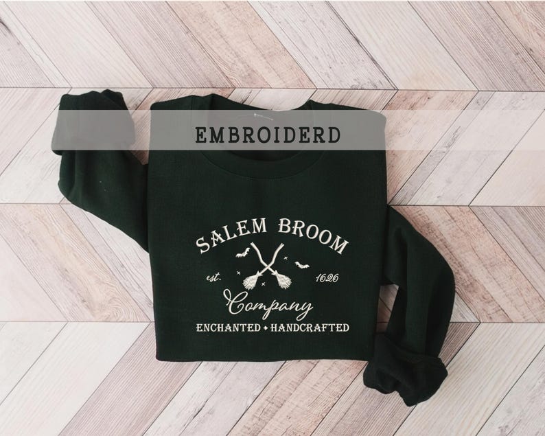 K&ouml;nnte beinhalten: Ein schwarzer Pullover mit wei&szlig; gesticktem Text, der "Salem Broom Company est. 1626 Enchanted + Handcrafted" lautet. Das Design zeigt zwei gekreuzte Besen mit einem Stern und Flederm&auml;usen.