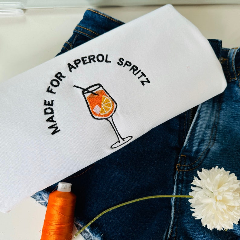 Puede incluir: Camiseta blanca con bordado negro que dice "Made for Aperol Spritz" y una imagen estilizada de un c&oacute;ctel Aperol Spritz.