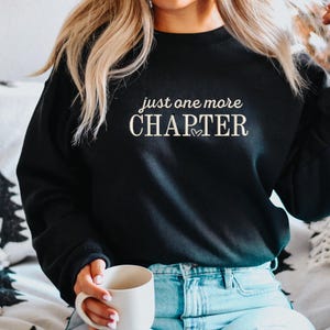 Pode incluir: Camisola preta com o texto "just one more CHAPTER" em escrita branca. Um pequeno cora&ccedil;&atilde;o est&aacute; entre as palavras. A camisola &eacute; usada com jeans azuis. Uma caneca branca &eacute; segurada na m&atilde;o.