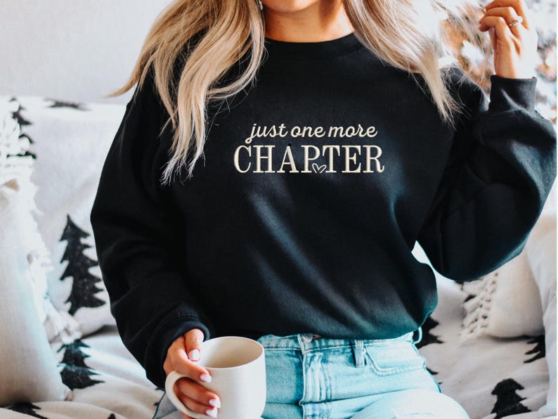 Pode incluir: Camisola preta com o texto "just one more CHAPTER" em escrita branca. Um pequeno cora&ccedil;&atilde;o est&aacute; entre as palavras. A camisola &eacute; usada com jeans azuis. Uma caneca branca &eacute; segurada na m&atilde;o.