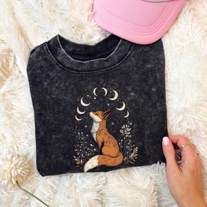 May include: A black t-shirt featuring a fox illustration with a crescent moon design. The fox is brown and white, surrounded by plants and moons. A pink trucker hat is visible in the image.