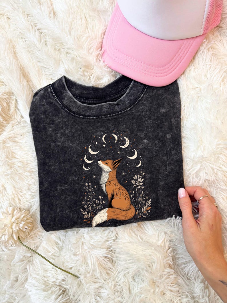 May include: A black t-shirt featuring a fox illustration with a crescent moon design. The fox is brown and white, surrounded by plants and moons. A pink trucker hat is visible in the image.