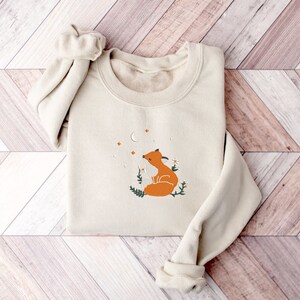 以下が含まれることがあります： 三日月と星の下、白い花畑に座っているオレンジ色のキツネのグラフィックが描かれたベージュのトレーナーです。