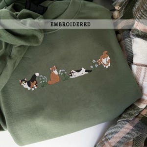 Può includere: Felpa verde oliva con ricamo di quattro gatti e fiori. I gatti sono raffigurati in varie pose, con motivi calico, arancioni e bianchi e neri. La parola "EMBROIDERED" è stampata in alto.