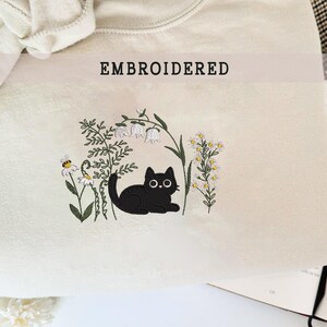 Könnte beinhalten: Cremefarbenes Sweatshirt mit einem gestickten Design mit einer schwarzen Katze, Gänseblümchen und anderem Grün. Das Wort "EMBROIDERED" ist oben aufgedruckt. Die Katze liegt und schaut nach vorne.