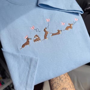 Könnte beinhalten: Hellblaues T-Shirt mit einer Stickerei von Dackeln, die mit rosa Schmetterlingen spielen. Die Dackel sind in verschiedenen Posen dargestellt und interagieren mit den Schmetterlingen. Ein verspieltes und skurriles Design.