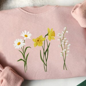 May include: A pink sweatshirt with a floral design featuring white daisies, yellow daffodils, and white lily of the valley flowers.
