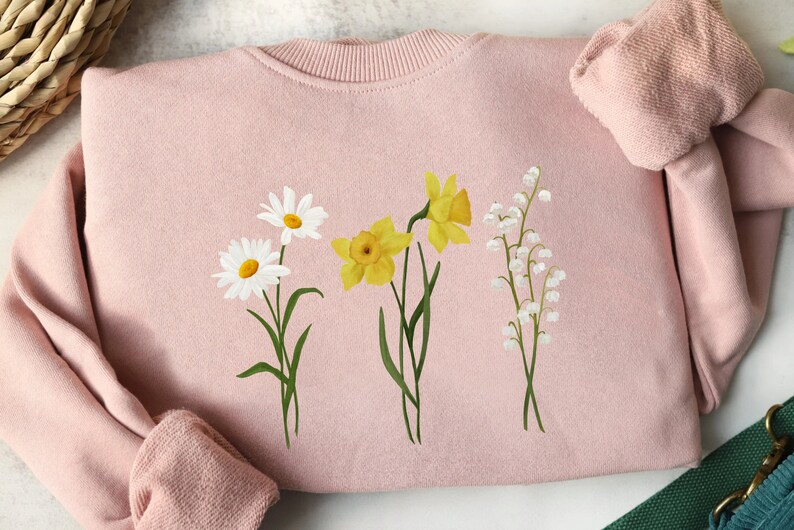 May include: A pink sweatshirt with a floral design featuring white daisies, yellow daffodils, and white lily of the valley flowers.