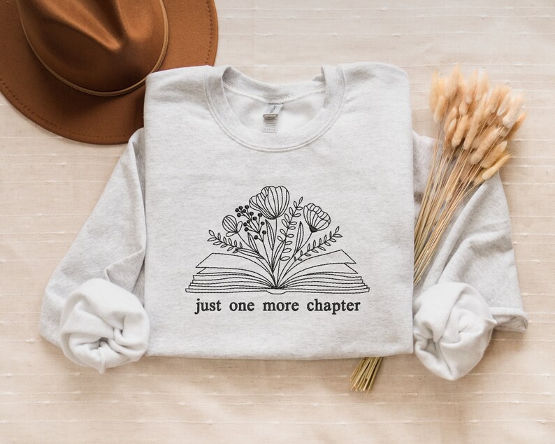 May include: A gray sweatshirt with a black and white illustration of an open book with flowers growing out of it. The text "just one more chapter" is printed below the illustration.