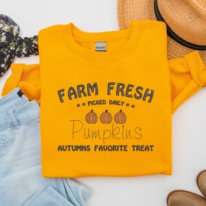 Op de afbeelding: Mosterdgeel sweatshirt met de tekst "FARM FRESH PUMPKINS" in zwart script. Het ontwerp bevat drie oranje pompoenen en de zin "AUTUMNS FAVORITE TREAT". Het sweatshirt is opgevouwen met een strohoed en jeans op de achtergrond.