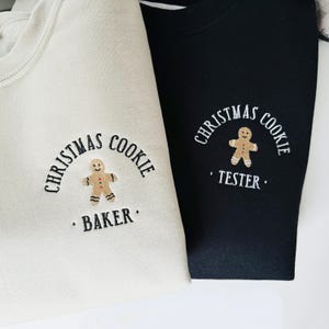 May include: Two sweatshirts, one cream and one black, featuring embroidered text and a gingerbread man design. The cream sweatshirt says "Christmas Cookie Baker" and the black one says "Christmas Cookie Tester". Both have a crew neck.