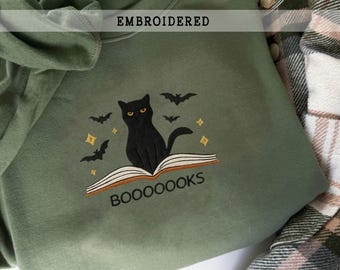 以下が含まれることがあります： オリーブグリーンのスウェットシャツ。刺繍のデザインは、開いた本の上に座っている黒猫と「BOOOOOKS」の文字が特徴です。デザインにはコウモリと星も含まれています。「EMBROIDERED」の文字が上部に印刷されています。