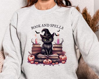 Könnte beinhalten: Hellgraues Sweatshirt mit Halloween-Motiv. Das Design zeigt eine schwarze Katze mit Hexenhut, die auf Büchern sitzt, mit Kürbissen und Kerzen. Der Text "BOOK AND SPELLS" befindet sich über der Katze.