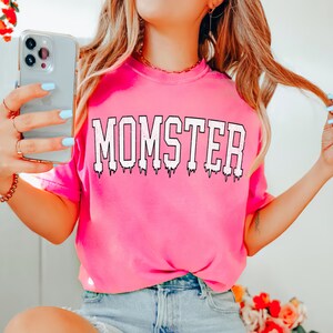 May include: Pink t-shirt with the word "MOMSTER" printed in white with a dripping effect.