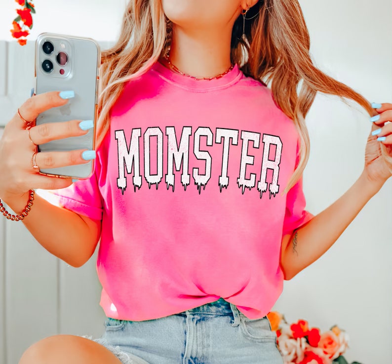 May include: Pink t-shirt with the word "MOMSTER" printed in white with a dripping effect.