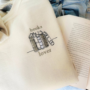 Puede incluir: Sudadera color crema con un diseño bordado de libros apilados y la palabra "books lover" en hilo gris. Un libro y unos vaqueros de mezclilla son visibles en el fondo.