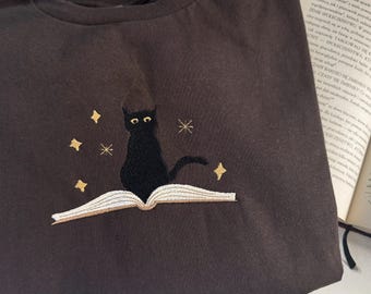 以下が含まれることがあります： 黒い猫が本の上に座っている刺繍のデザインが施された濃い茶色のTシャツ。猫の周りに金色の星が散りばめられています。
