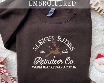 May include: A dark brown embroidered sweatshirt with the text "EMBROIDERED" across the top. The sweatshirt features an embroidered design with the words "SLEIGH RIDES", "Reindeer Co.", and "WARM BLANKETS AND COCOA".
