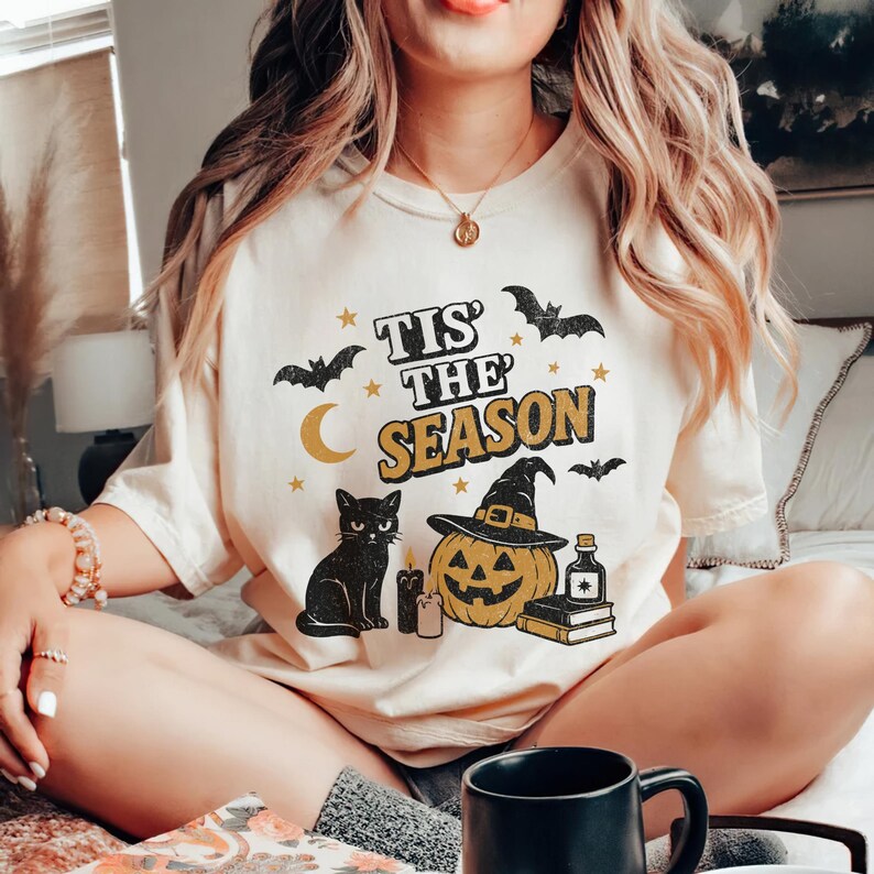 May include: Cream-colored t-shirt with a Halloween-themed graphic. The design includes the text "Tis' The Season", a black cat, a jack-o'-lantern wearing a witch hat, bats, stars, a crescent moon, candles, a potion bottle, and books.
