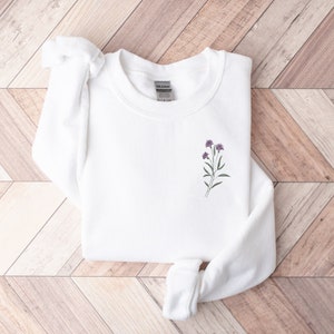 May include: White long-sleeved sweatshirt with a small purple flower embroidery on the front.