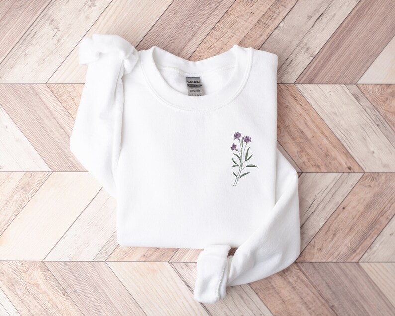 May include: White long-sleeved sweatshirt with a small purple flower embroidery on the front.