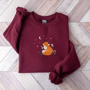 以下が含まれることがあります： 三日月と星を見上げるキツネの刺繍が施された、マルーンのスウェットシャツ。キツネはオレンジと白で、デザインは小さな白い花で囲まれています。