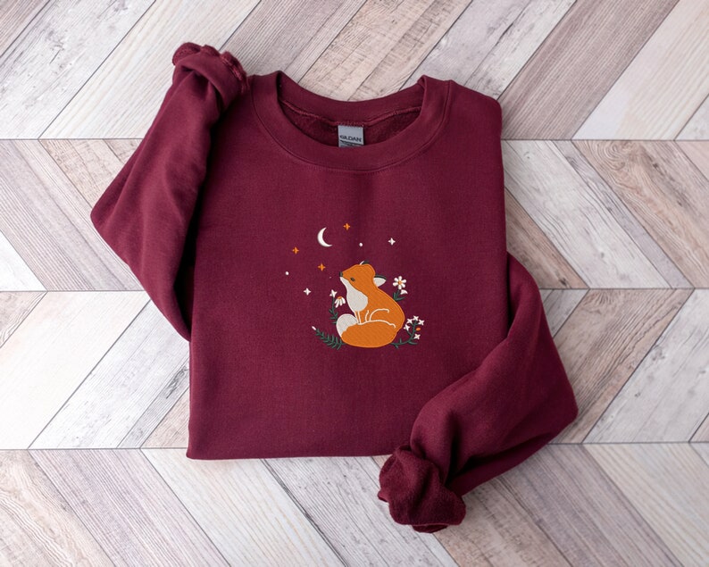 以下が含まれることがあります： 三日月と星を見上げるキツネの刺繍が施された、マルーンのスウェットシャツ。キツネはオレンジと白で、デザインは小さな白い花で囲まれています。