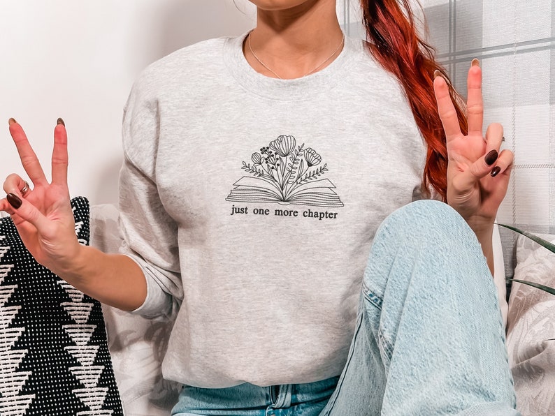 May include: Light gray sweatshirt with a floral book design and the text "just one more chapter" in black. The design features an open book with flowers and leaves growing from it. The sweatshirt is a crew neck style.