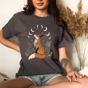 K&ouml;nnte beinhalten: Dunkelgraues T-Shirt mit einer Illustration eines Fuchses, der zu Halbmonden und Sternen aufblickt. Der Fuchs ist braun und wei&szlig; dargestellt, umgeben von floralen Details. Das Design ist mittig auf dem T-Shirt platziert.