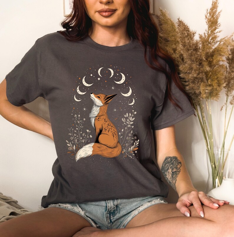 K&ouml;nnte beinhalten: Dunkelgraues T-Shirt mit einer Illustration eines Fuchses, der zu Halbmonden und Sternen aufblickt. Der Fuchs ist braun und wei&szlig; dargestellt, umgeben von floralen Details. Das Design ist mittig auf dem T-Shirt platziert.