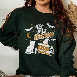 May include: Dark green sweatshirt with a Halloween-themed graphic. The design includes a white cat, a jack-o'-lantern wearing a witch hat, bats, stars, a crescent moon, and the text "Tis the Season."