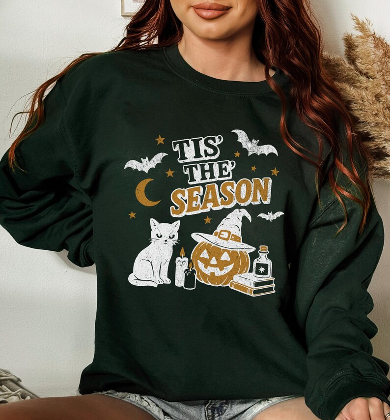 May include: Dark green sweatshirt with a Halloween-themed graphic. The design includes a white cat, a jack-o'-lantern wearing a witch hat, bats, stars, a crescent moon, and the text "Tis the Season."