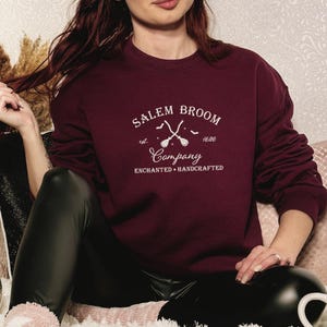 K&ouml;nnte beinhalten: Ein maroonfarbener Pullover mit einem wei&szlig;en Grafikdesign, auf dem "Salem Broom Company est. 1626 Enchanted Handcrafted" steht. Die Grafik zeigt auch zwei gekreuzte Besen mit Flederm&auml;usen, die um sie herum fliegen.