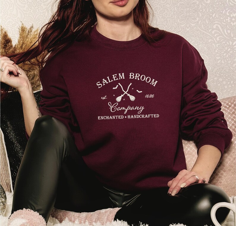 K&ouml;nnte beinhalten: Ein maroonfarbener Pullover mit einem wei&szlig;en Grafikdesign, auf dem "Salem Broom Company est. 1626 Enchanted Handcrafted" steht. Die Grafik zeigt auch zwei gekreuzte Besen mit Flederm&auml;usen, die um sie herum fliegen.