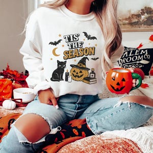 May include: White sweatshirt with a Halloween-themed graphic. The design includes a black cat, a jack-o'-lantern wearing a witch hat, bats, stars, and the text "Tis the Season". A person is holding an orange pumpkin mug.