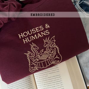 Puede incluir: Sudadera burdeos con el texto bordado "HOUSES & HUMANS" y un gráfico de tres dragones jugando a un juego de mesa. La palabra "EMBROIDERED" está encima del texto principal.