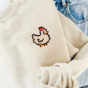 Puede incluir: Una sudadera beige con un diseño de pollo bordado en píxeles. El pollo es blanco con una cresta roja y patas marrones.