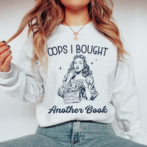 Può includere: Felpa grigio chiaro con la scritta "OOPS I BOUGHT Another Book" sopra un'illustrazione in bianco e nero di una donna che tiene dei libri. La felpa è a girocollo.