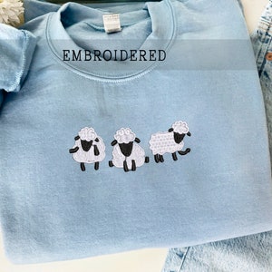 May include: A light blue sweatshirt with the word "EMBROIDERED" in black text. Three white sheep with black faces are embroidered on the front of the sweatshirt.