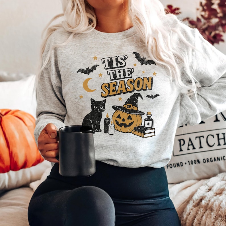 May include: A light gray sweatshirt with a Halloween-themed graphic. The design includes a black cat, a jack-o'-lantern wearing a witch hat, bats, stars, and the text "Tis the Season". A black mug is held in the hand.