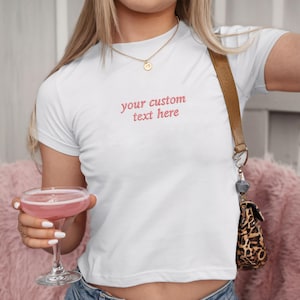Może przedstawiać: Biały t-shirt z wyhaftowanym napisem "your custom text here" w kolorze czerwonym. Koszulka jest z krótkim rękawem i noszona ze złotym naszyjnikiem i brązową torbą na ramię. Różowy koktajl trzymany jest w przezroczystym szkle.