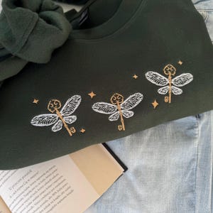 Op de afbeelding: Een donkergroen sweatshirt met drie geborduurde witte sleutel-vormige libellen met gouden accenten. De libellen zijn omringd door kleine gouden sterren.