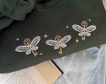 May include: A dark green sweatshirt with three embroidered white key-shaped dragonflies with gold accents. The dragonflies are surrounded by small gold stars.