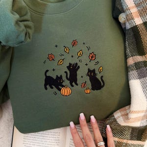 May include: An olive green crewneck sweatshirt featuring an embroidered design of three black cats playing with orange pumpkins and falling autumn leaves in shades of yellow, orange, and brown, with small star accents surrounding the cats.