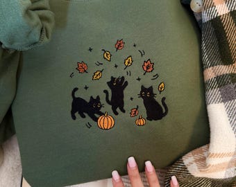 May include: An olive green crewneck sweatshirt featuring an embroidered design of three black cats playing with orange pumpkins and falling autumn leaves in shades of yellow, orange, and brown, with small star accents surrounding the cats.