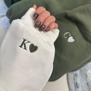 Op de afbeelding: Close-up van twee sweatshirts, één wit en één olijfgroen. De witte manchet van de sweatshirt heeft de letter "K" en een groen hart geborduurd. De olijfgroene sweatshirtmanchet heeft de letters "C" en een wit hart geborduurd.