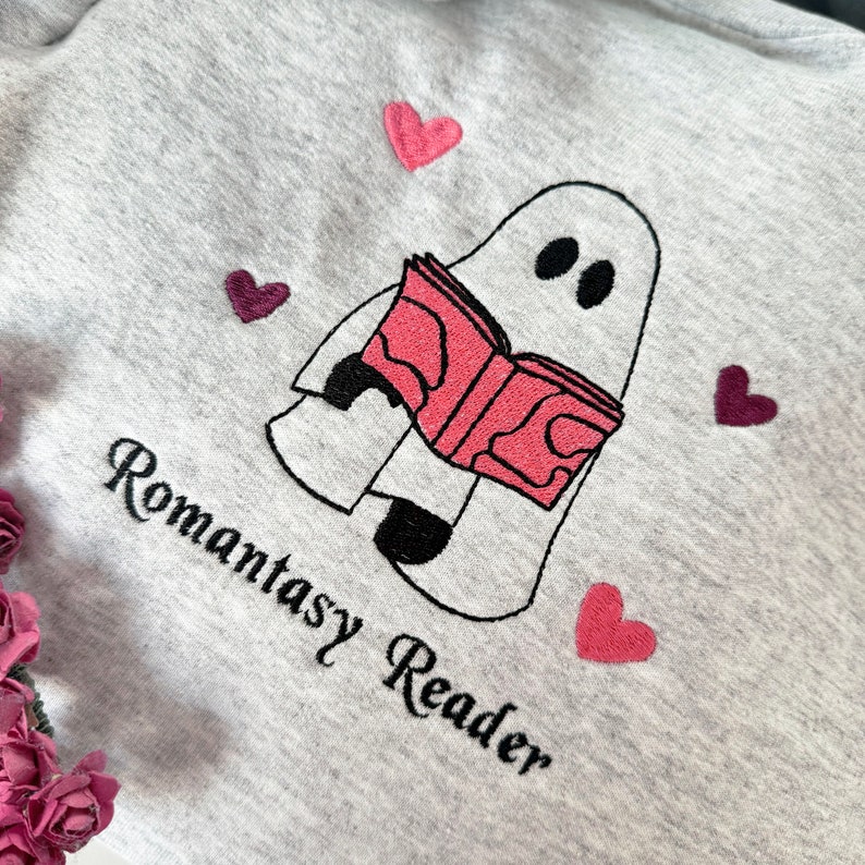 May include: A gray sweatshirt with a white ghost reading a pink book. The ghost has two black eyes. There are pink and purple hearts around the ghost. The text "Romantasy Reader" is embroidered on the sweatshirt.