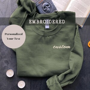 Può includere: Una felpa verde scuro con la parola "EMBROIDERED" stampata in nero in alto e la parola "custom" ricamata in bianco sul davanti. La felpa è piegata e appoggiata su una superficie scura.